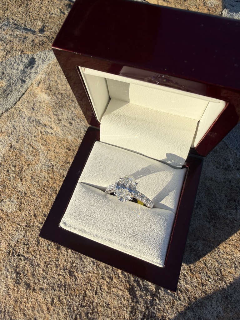 Oval-cut lab-grown diamond engagement ring with a diamond-set band in white gold, in a dark wood box with cream velvet interior, photographed on stone in sunlight