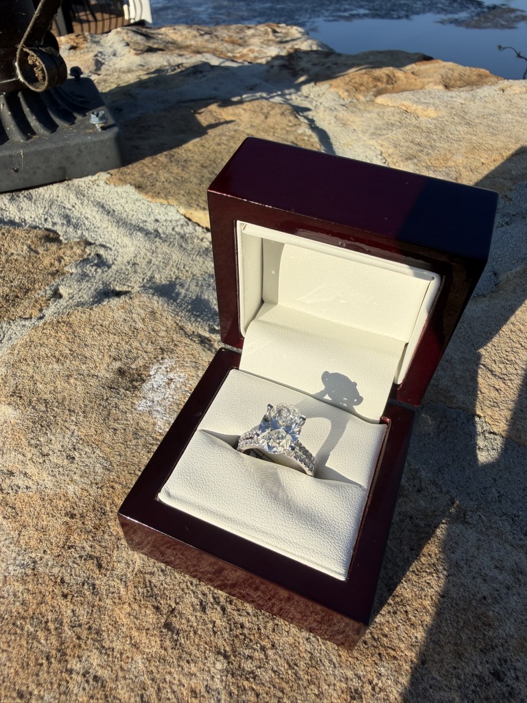 Oval-cut lab-grown diamond bridal set with diamond-set band in white gold, in a dark wood box with cream velvet interior, photographed on stone near water in sunlight