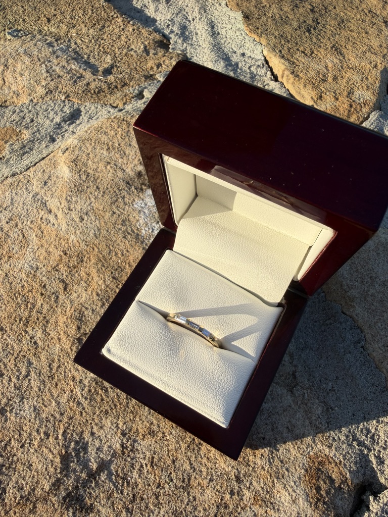Matching yellow gold diamond wedding band with baguette stones, in a deep red wood box with cream velvet interior, photographed on stone in sunlight