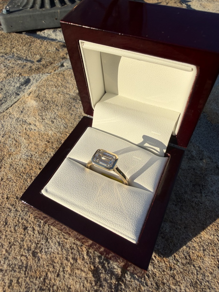 Emerald-cut lab-grown diamond solitaire with gold bezel in a deep red wood box with cream velvet interior, photographed on stone in sunlight