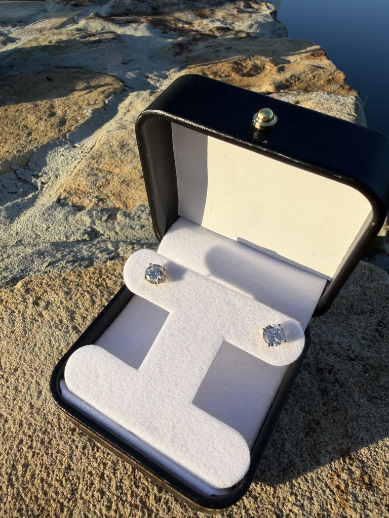 Pair of round lab-grown diamond stud earrings in yellow gold, in an open black leather box with white velvet inserts, photographed on stone near water