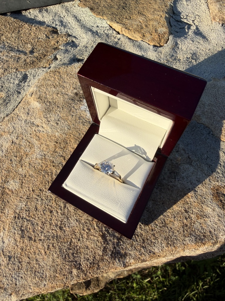 Round brilliant-cut lab-grown diamond solitaire engagement ring with tapered baguette side stones in yellow gold, in a dark wood box with cream velvet interior, photographed on stone in sunlight