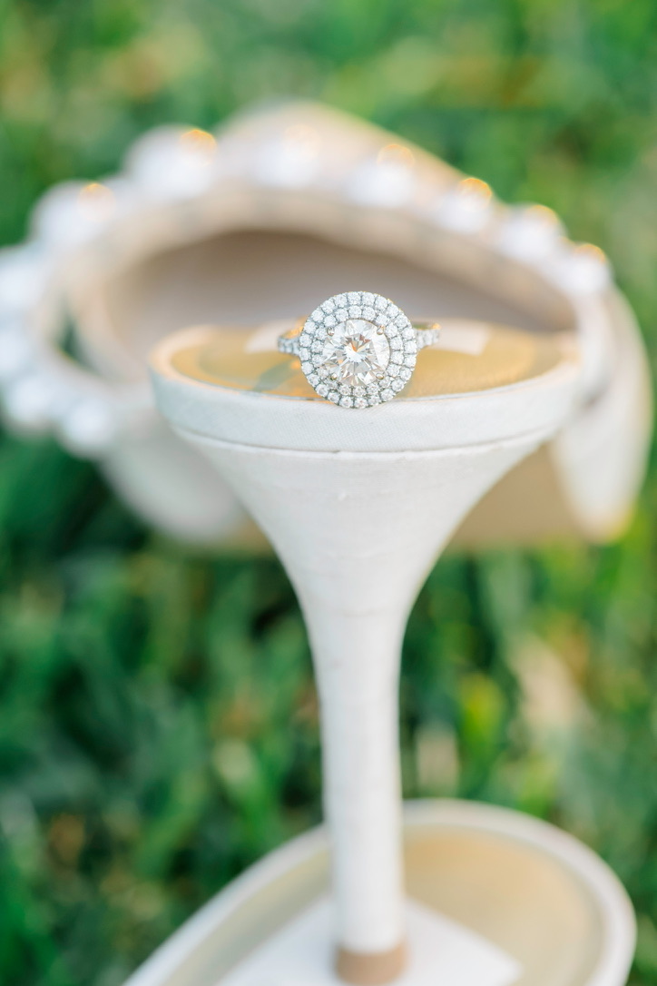 Double halo round brilliant engagement ring resting on the heel of a pearl-adorned bridal shoe in the grass