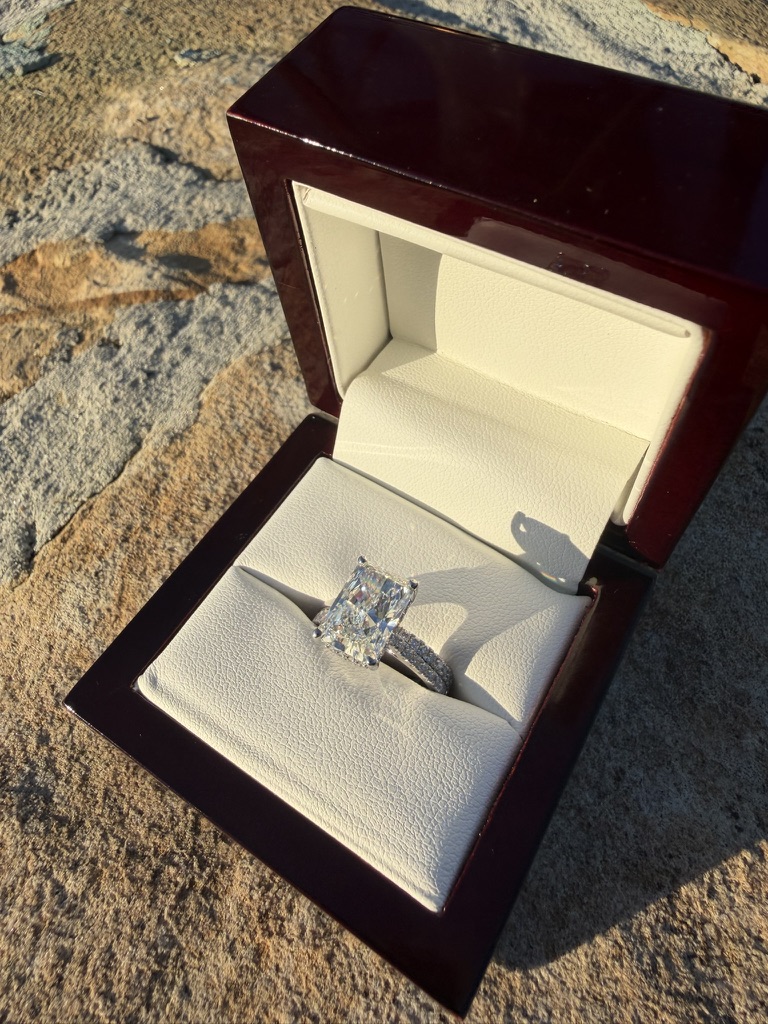 Radiant-cut lab-grown diamond bridal set with diamond-set band in white gold, in a dark wood box with cream velvet interior, photographed on stone in sunlight