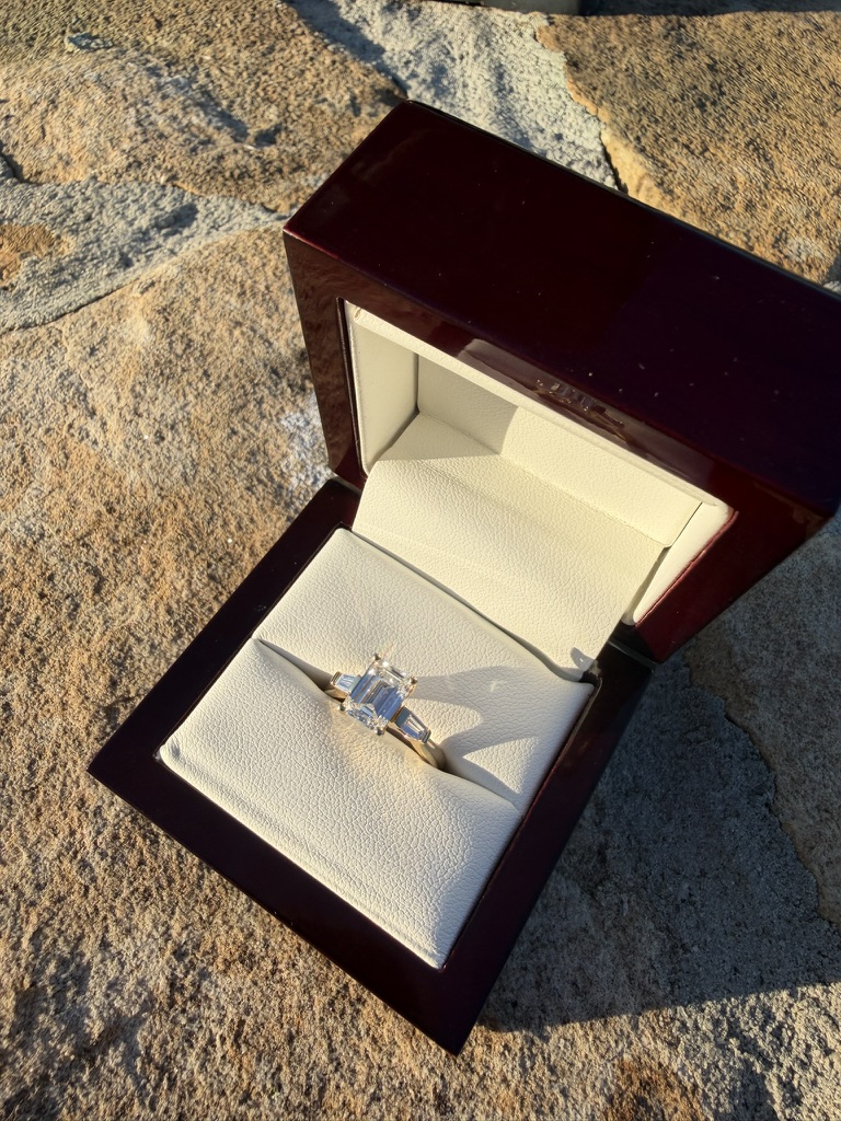 Emerald-cut lab-grown diamond engagement ring with tapered baguette side stones in yellow gold, in a deep red wood box with cream velvet interior, photographed on stone in sunlight