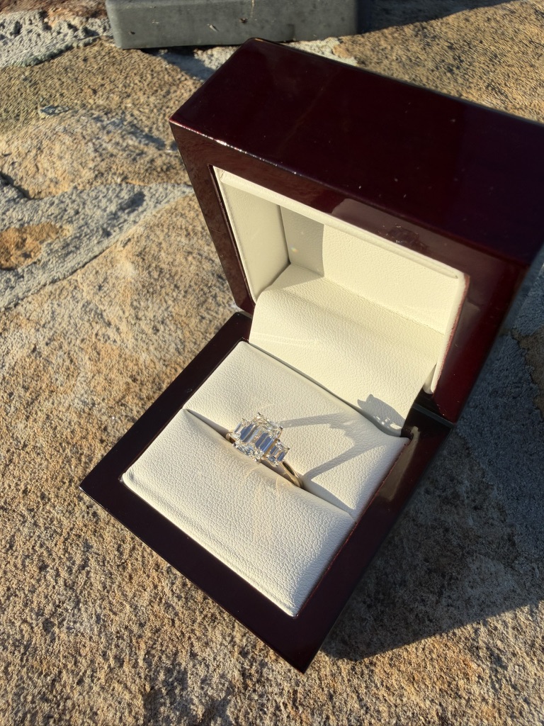 Three-stone emerald-cut lab-grown diamond ring in yellow gold with tapered baguette side stones, in a deep red wood box with cream velvet interior, photographed on stone in sunlight