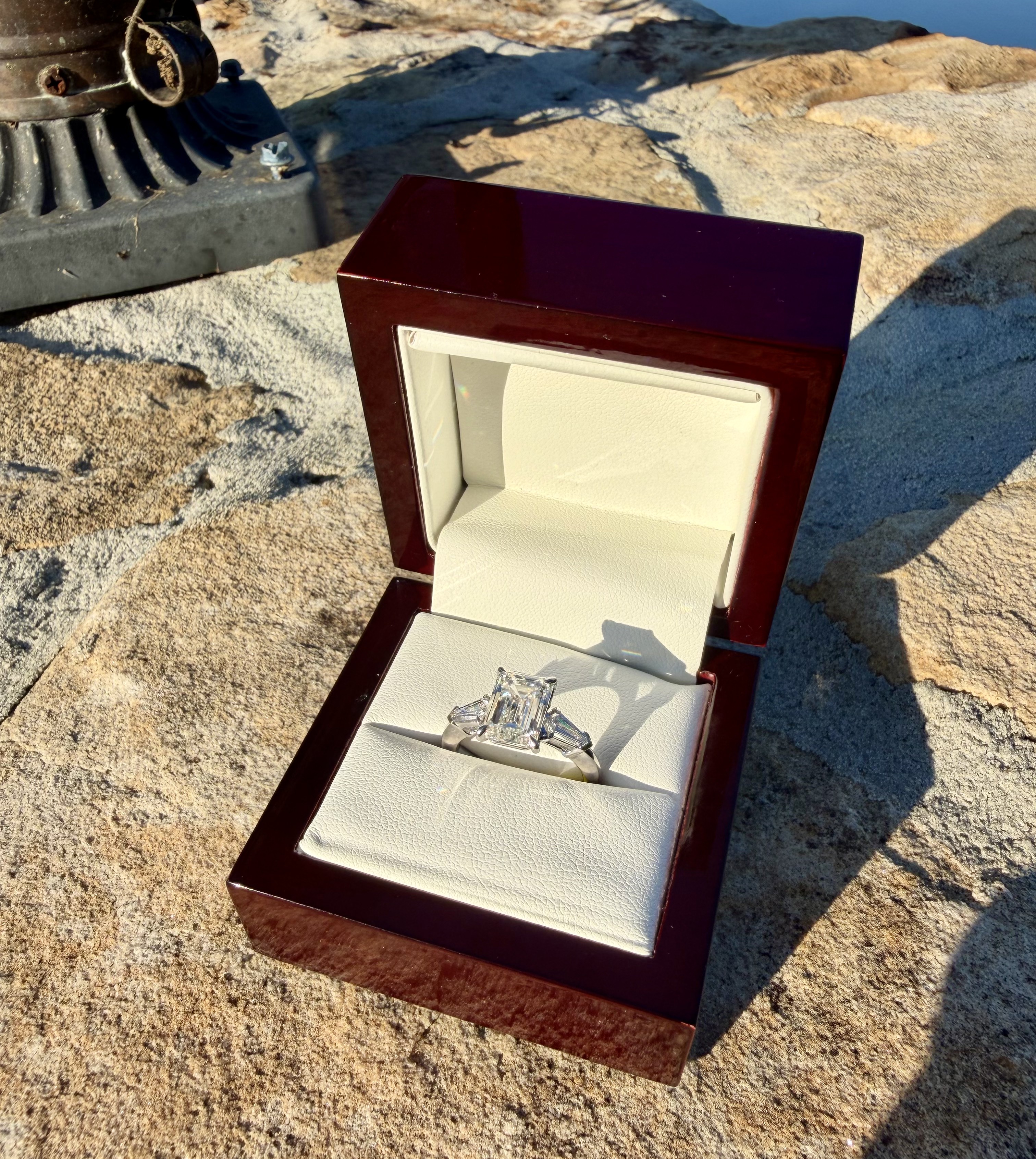 Emerald-cut lab-grown diamond engagement ring in white gold with tapered baguette side stones, in its glossy dark cherry wood box with cream leather interior, photographed on stone in bright sunlight