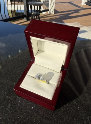 Round brilliant lab-grown diamond engagement ring with a yellow-gold center setting on a white-gold band, standing upright in a glossy dark cherry wood box with cream leather interior, photographed on a stone table outdoors