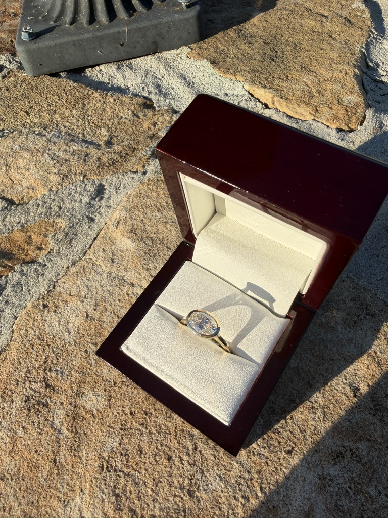Oval lab-grown diamond engagement ring in a deep red wood box with cream velvet interior, photographed on stone in sunlight