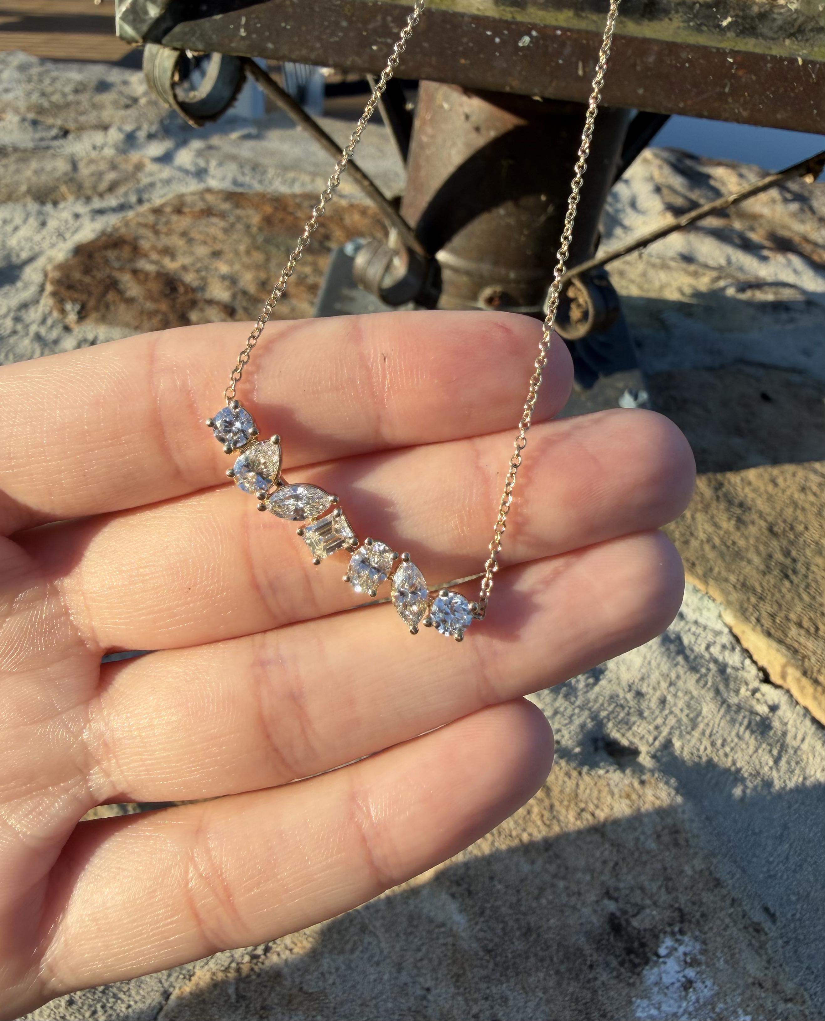 Lab-grown diamond necklace with graduated mixed-shape stones — round, pear, marquise, emerald cut and oval — on a fine yellow gold chain, held in a hand outdoors