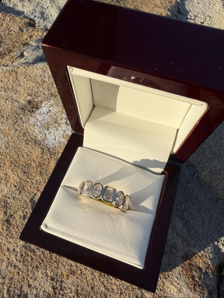 Bezel-set lab-grown diamond band with mixed oval and emerald-cut stones in white gold, in a deep red wood box with cream velvet interior, photographed on stone in sunlight