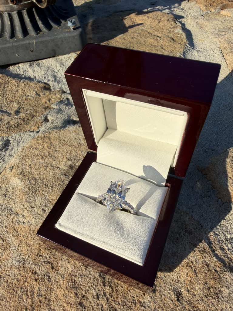 Marquise-cut lab-grown diamond engagement ring in white gold with a diamond-set band, standing upright in a glossy dark cherry wood box with cream leather interior, photographed on stone in sunlight