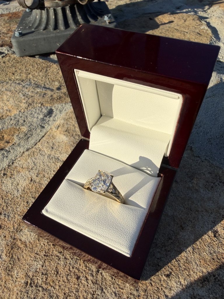 Elongated cushion-cut lab-grown diamond engagement ring with diamond band in yellow gold, in a deep red wood box with cream velvet interior, photographed on stone in sunlight