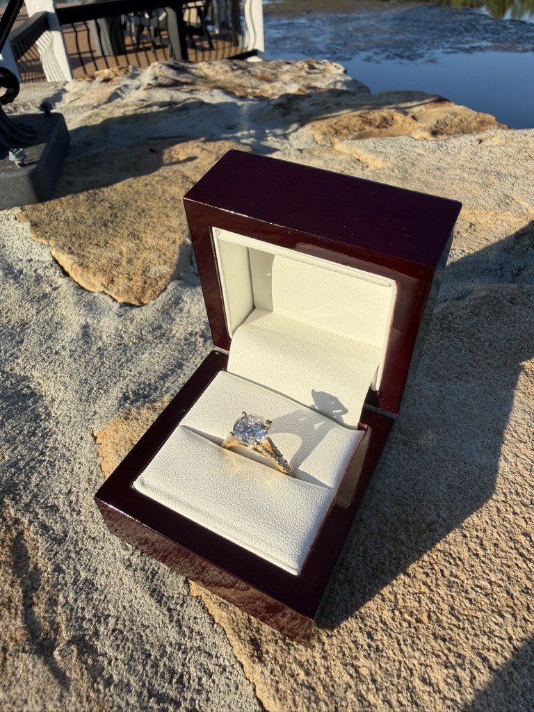 Round brilliant-cut lab-grown diamond engagement ring with diamond-set band in yellow gold, in a deep red wood box with cream velvet interior, photographed on stone near water in sunlight