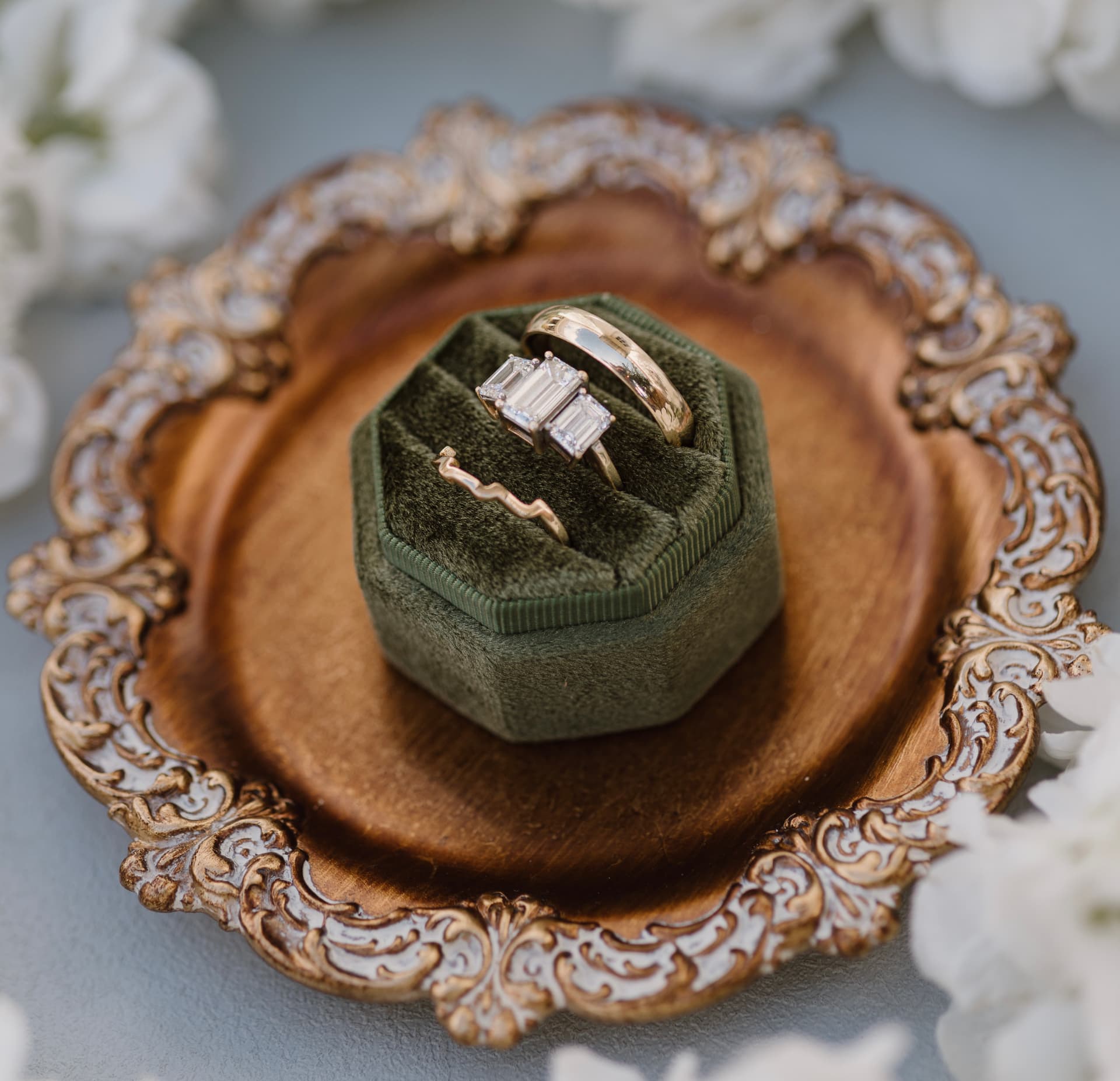 Emerald-cut engagement ring set and gold wedding bands on a green velvet box atop an antique gold-rimmed tray with white flowers