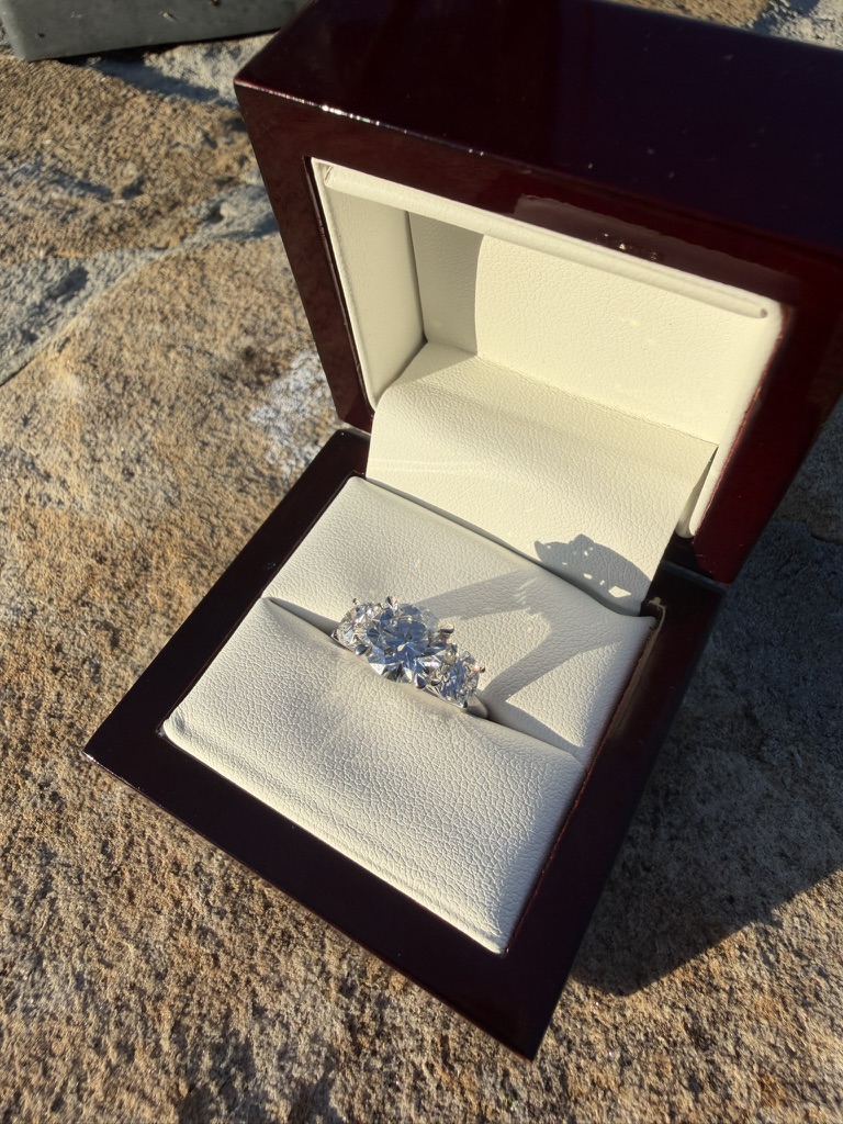 Three-stone round brilliant engagement ring in a dark wood box with cream velvet interior, photographed outdoors in sunlight
