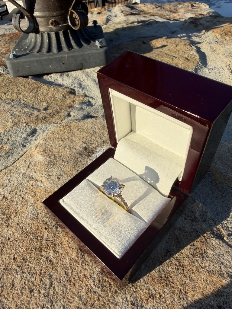 Large round brilliant lab-grown diamond engagement ring in yellow gold with a pavé band, in its glossy dark cherry wood box with cream leather interior, photographed on stone in bright sunlight