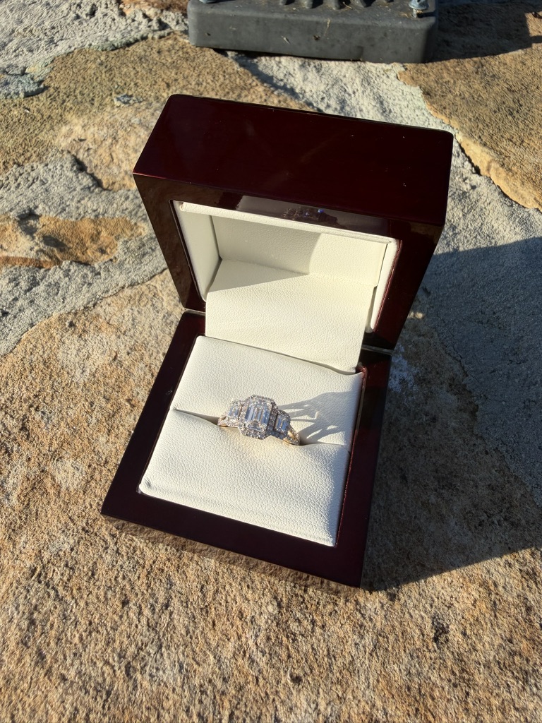 Emerald-cut lab-grown diamond engagement ring with tapered baguette side stones in yellow gold, in a deep red wood box with cream velvet interior, photographed on stone in sunlight